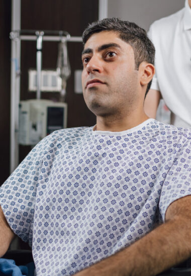 Serious male hospital patient in patterned gown sits in bed, gazing to the side in a clinical setting.