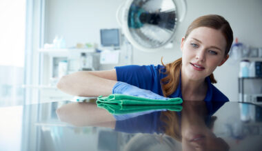Young smiling woman in blue scrubs dusting hospital room mirror with green cloth