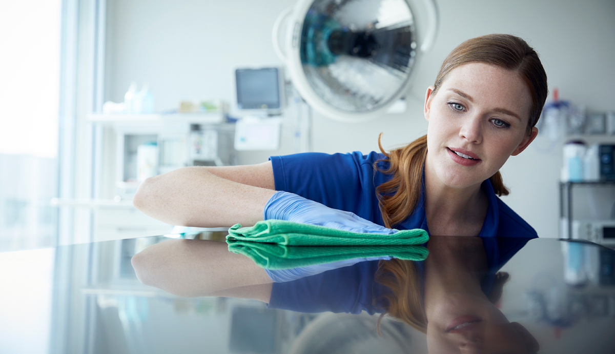 Young smiling woman in blue scrubs dusting hospital room mirror with green cloth