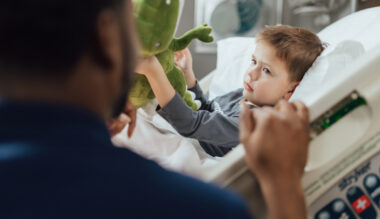 Adorable toddler boy curiously reaches for stuffed green dinosaur toy held by unidentified person.