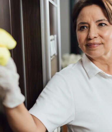 Smiling mature Latina housekeeper cleaning window in healthcare facility, wearing white polo shirt.