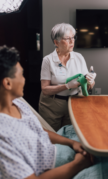 An elderly woman in a hospital gown sits at a table while a nurse holds a spirometer, assisting with a breathing exercise.