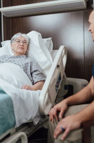 Elderly patient in hospital bed speaks with healthcare worker, receiving compassionate care.