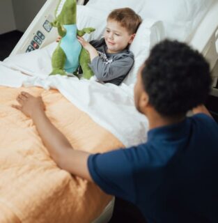 The image shows a young child playing with plush toys on a bed while an adult sits nearby and watches over them attentively. The child has a delighted expression as they handle the plush dinosaur toy. The scene conveys a sense of comfort, safety and engagement between the adult caregiver and happy child.
