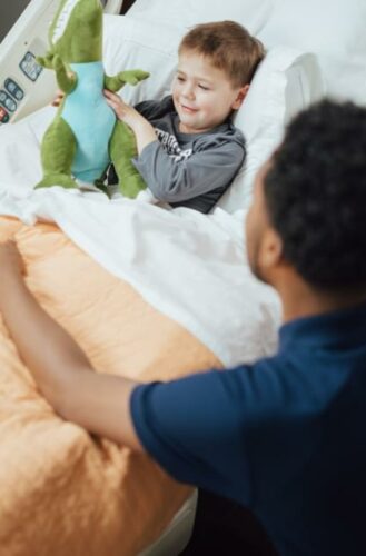 The image shows a young child playing with plush toys on a bed while an adult sits nearby and watches over them attentively. The child has a delighted expression as they handle the plush dinosaur toy. The scene conveys a sense of comfort, safety and engagement between the adult caregiver and happy child.