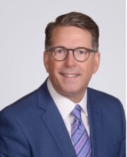 Professional headshot of a smiling middle-aged man in glasses, blue suit and striped tie on gray background