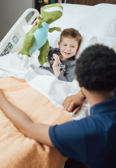 A young child smiles while playing with a green stuffed animal in a hospital bed as an adult looks on.