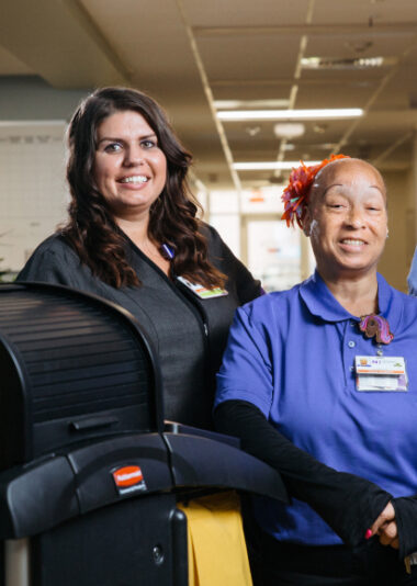 Smiling Crothall Healthcare employees in uniform inside a building, one seated and one standing nearby.