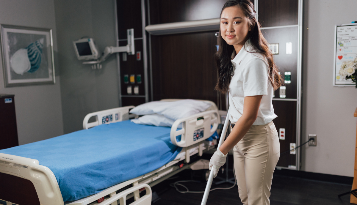 Alt text: Smiling healthcare worker in uniform standing next to hospital bed in patient room