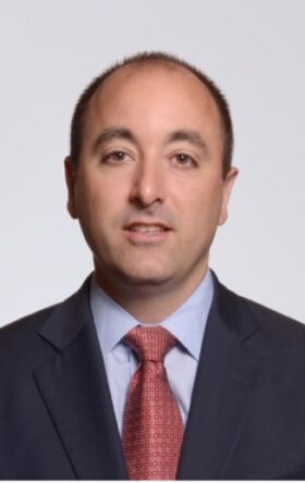 Middle-aged businessman in navy suit and red tie smiling at camera against gray background.
