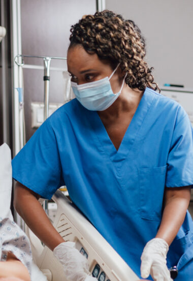 Nurse in blue scrubs and face mask looking down while adjusting hospital bed controls.
