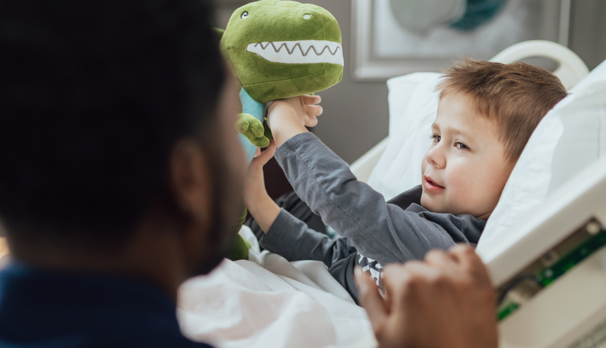 Young boy holding a green dinosaur stuffed animal while being carried by an adult