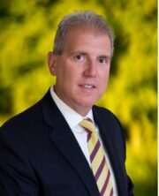 Middle-aged businessman with gray hair wearing navy suit and striped tie smiling in front of blurred green background