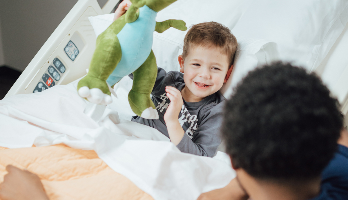 Young boy smiling and playing with stuffed animal in hospital bed, interacting with person out of frame