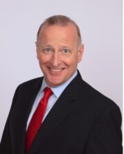 Smiling businessman with short grey hair wearing a dark suit, light blue shirt and red tie against a white background.