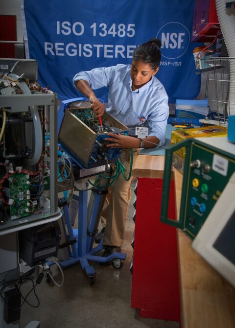 Student technician repairs medical equipment in ISO 13485 registered lab at NSF-ISR certification facility.