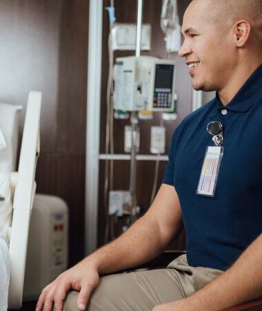 Smiling healthcare worker sitting in hospital room, wearing ID badge and blue polo shirt