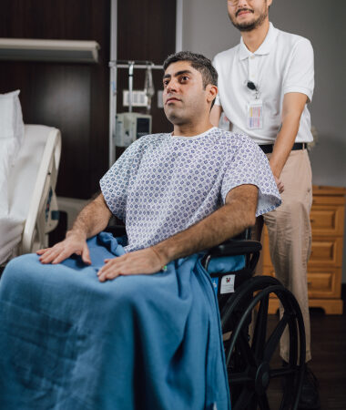 Patient in hospital gown in wheelchair with healthcare worker standing behind in patient room