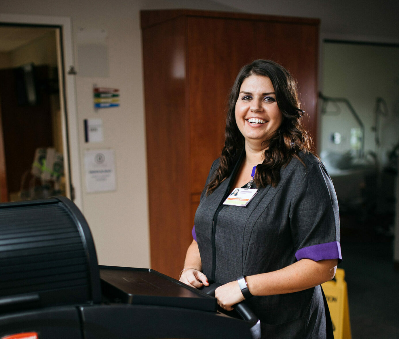 Smiling healthcare worker holding clipboard at nurse's station, wearing ID badge