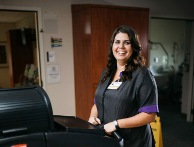 Smiling healthcare worker holding clipboard at nurse's station, wearing ID badge