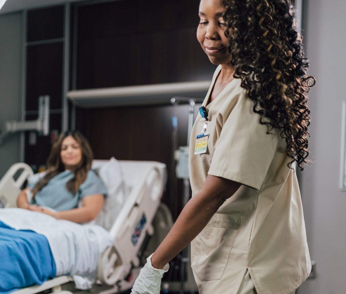 Curly-haired nurse in scrubs smiles while visiting a patient in a hospital room