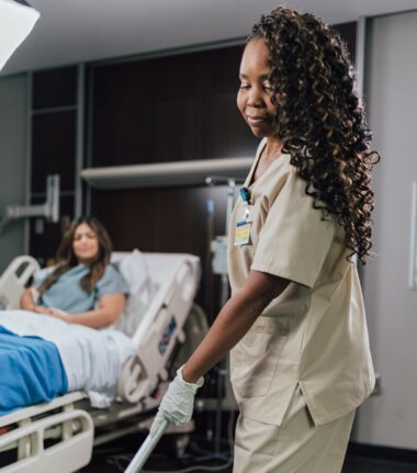Curly-haired nurse in scrubs smiles while visiting a patient in a hospital room
