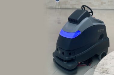 Autonomous floor cleaning robot with blue lights on gray tiled floor in commercial space.