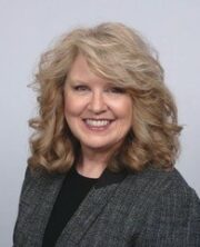 Smiling blonde woman with curly hair wearing a gray blazer, professional headshot on white background