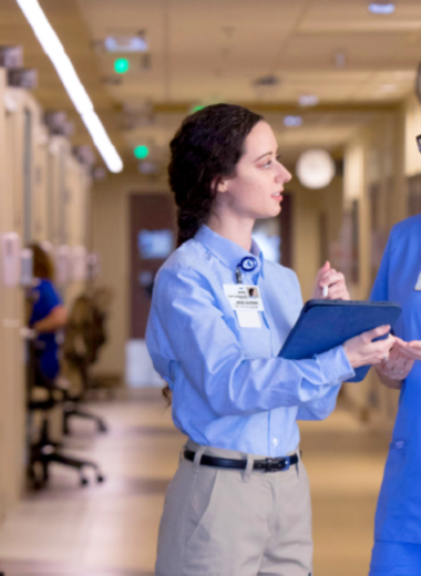 Two healthcare workers in blue scrubs having a discussion in a hospital hallway, holding clipboards.
