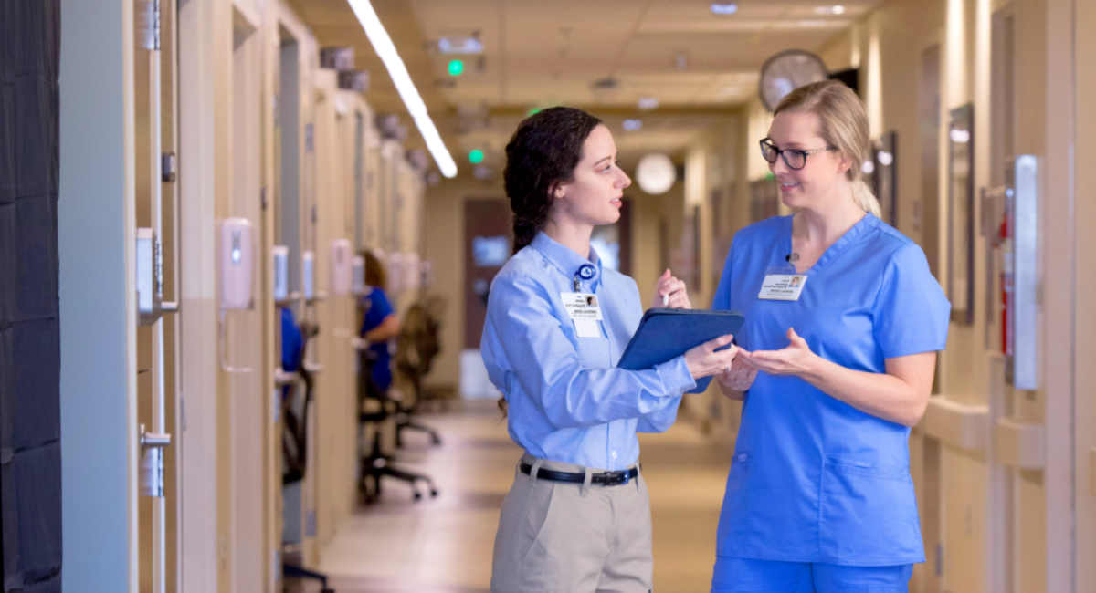 Two healthcare workers in blue scrubs having a discussion in a hospital hallway, holding clipboards.