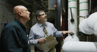Two men inspecting machinery in an industrial setting with large tanks and pipes.
