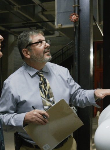 Two men inspecting machinery in an industrial setting with large tanks and pipes.