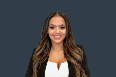 Smiling woman with long brown hair wearing a black blazer and white top against a dark blue background