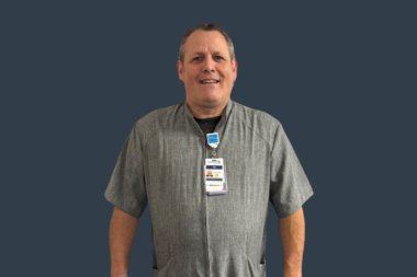 Smiling Crothall Healthcare employee wearing a grey shirt and ID badge on a dark blue background.