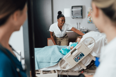 Smiling nurse adjusting baby in hospital bassinet as colleagues converse, healthcare support services