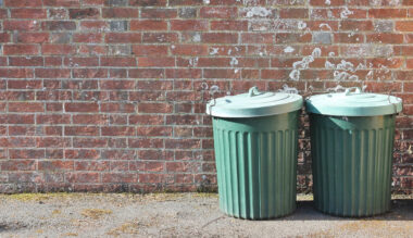 Green plastic garbage bins against a red brick wall with graffiti and gravel ground.