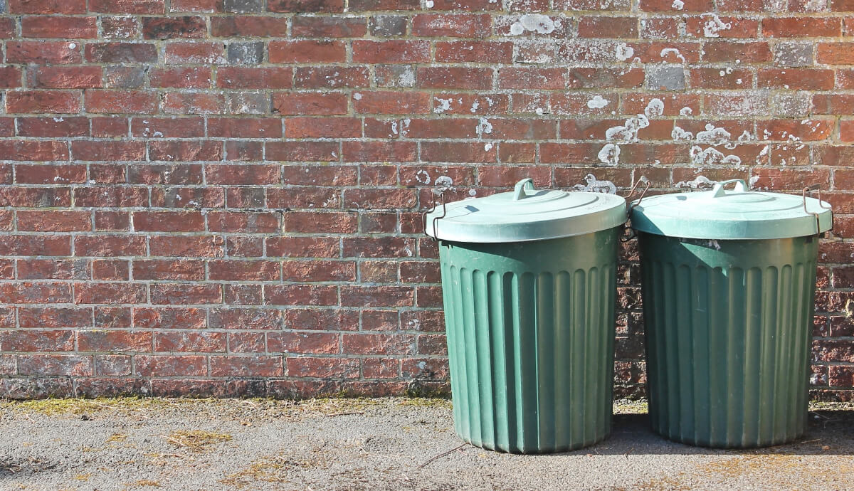 Green plastic garbage bins against a red brick wall with graffiti and gravel ground.