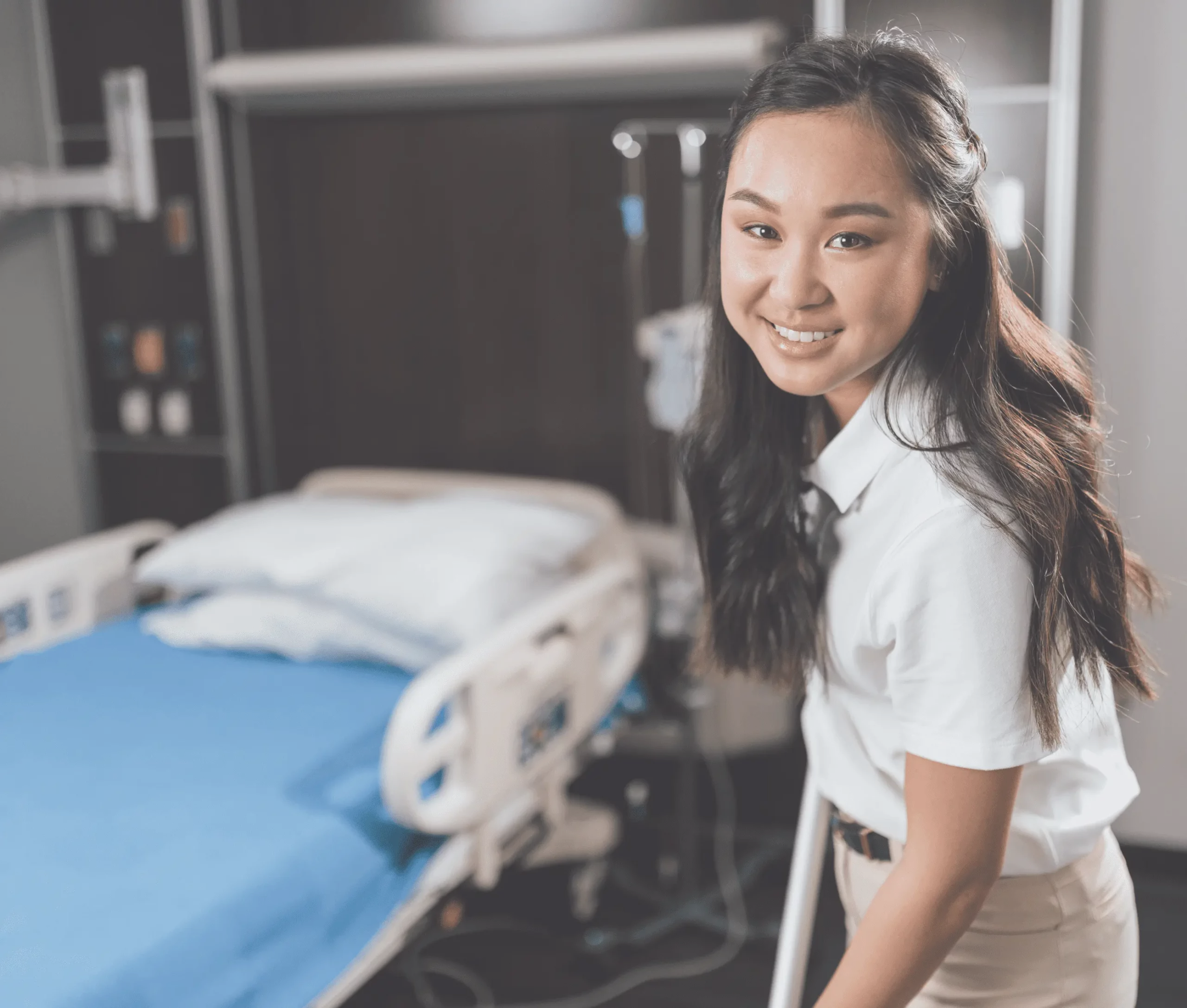 Smiling female healthcare worker standing near hospital bed, providing friendly patient care.