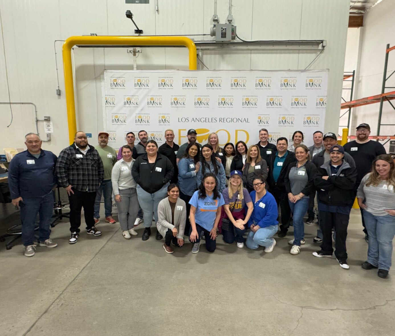 A group of volunteers poses at the Los Angeles Regional Food Bank, smiling in a warehouse setting with the food bank's logo in the background.