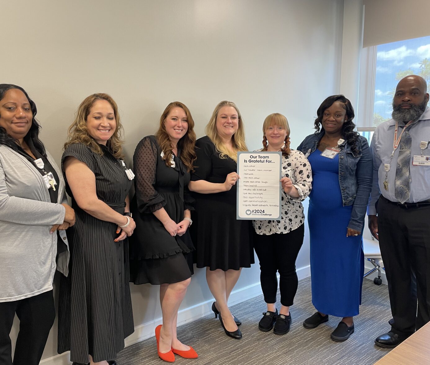 A group of seven professionals poses in an office, smiling while holding a sign that lists things their team is grateful for. They are dressed in business attire, with ID badges visible.